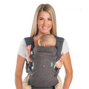 A woman with blonde hair, wearing a white t-shirt and khaki pants, smiling as she wears a dark grey fabric baby carrier with a baby inside, facing outwards.