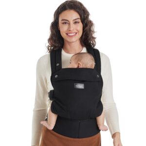 A smiling woman in a cream-colored sweater wearing a black baby carrier with a baby inside, facing outwards. The carrier is positioned on her front.