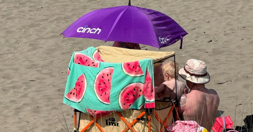 Best Baby Beach Gear for Sun Protection (2026): Tents, Rash Guards, Hats, and Sunscreen Family enjoying a sunny beach day with a colorful umbrella and watermelon towel.